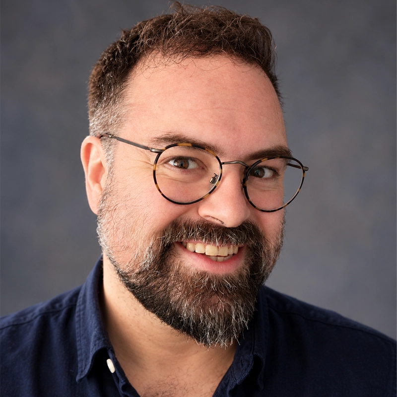 Danny Weller is a middle-aged man with a full beard and short, dark hair who smiles at the camera while wearing tortoiseshell glasses and a dark blue collared shirt against a textured, dark gray studio backdrop.