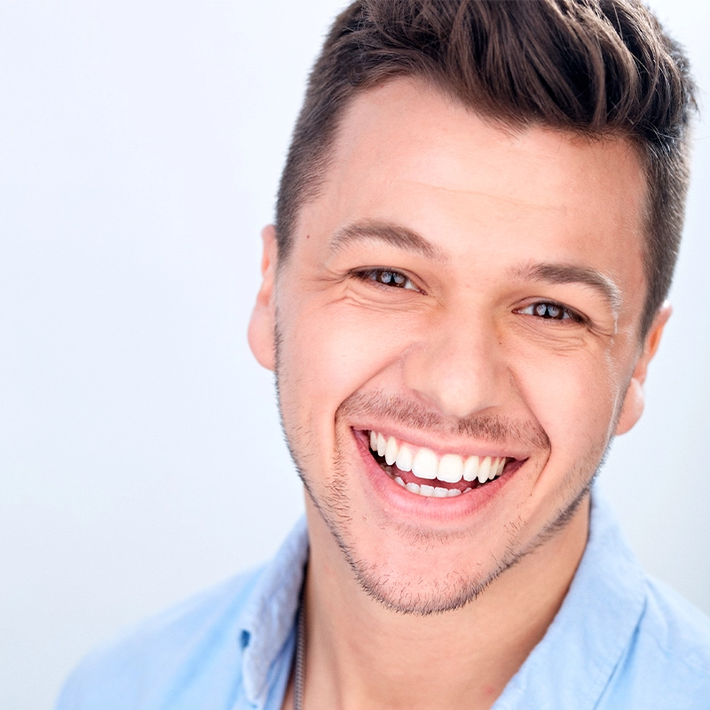 A young man with short brown hair and light stubble smiles broadly, showing his teeth. Wearing a light blue collared shirt, he stands against a plain background. The image is brightly lit, emphasizing his friendly expression, reminiscent of an Eve Tjoumakaris portrait.