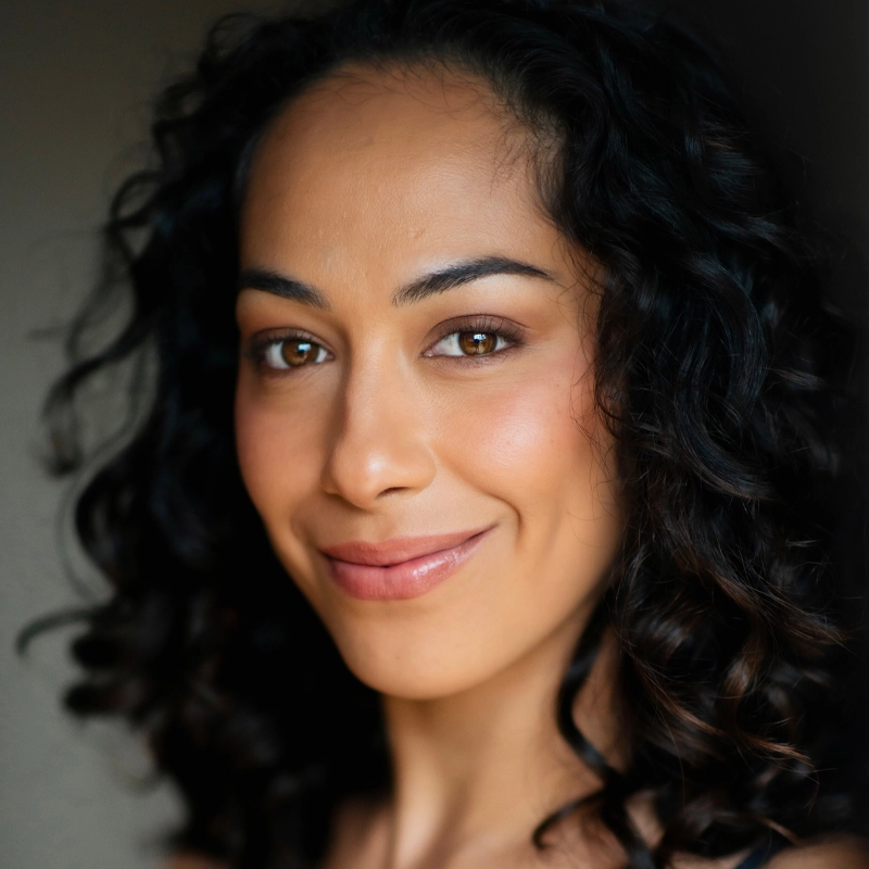 Heather Makalani, with medium skin tone and dark, curly hair, smiles softly. She has brown eyes, groomed eyebrows, and wears natural-looking makeup. The softly blurred background draws focus to her friendly, confident expression.