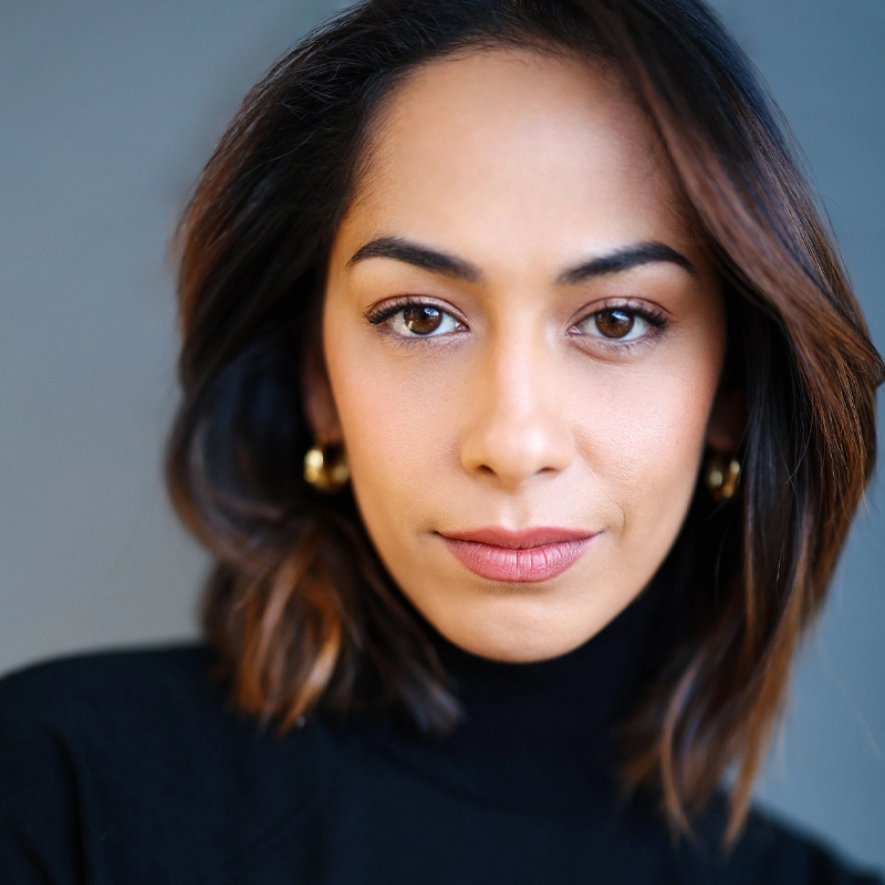 A woman with medium skin tone and shoulder-length brown hair looks directly at the camera with a neutral expression. Heather Makalani wears gold hoop earrings and a black turtleneck. The background is softly blurred in shades of blue and gray.