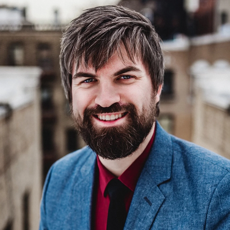 Danny Weller smiles at the camera, sporting a thick, dark beard and textured brown hair. He is wearing a blue blazer over a maroon shirt and a black necktie, with a blurred cityscape visible in the background.