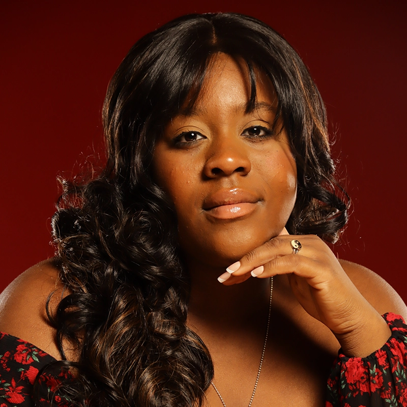 A woman with long, wavy dark hair and glowing skin poses against a deep red background. She gazes at the camera with a soft smile, resting her chin on her hand. She wears a floral off-shoulder top, a ring, and a necklace.