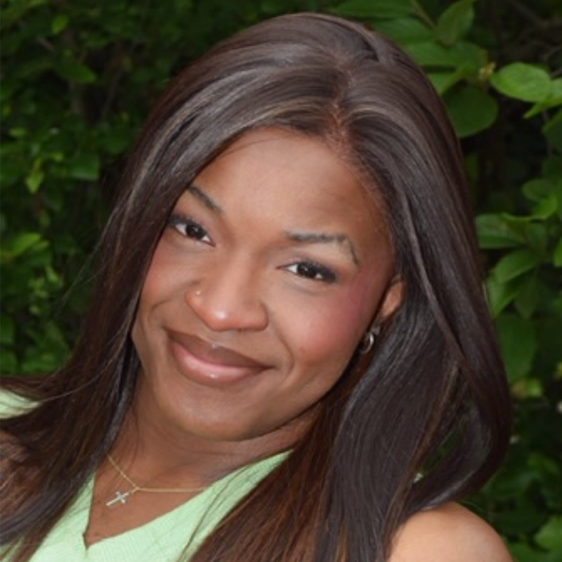 Nesziah Dennis, a woman with long, straight brown hair, smiles outdoors. She wears a light green top, a delicate necklace with a cross pendant, small earrings, and subtle makeup. Green leafy foliage fills the blurred background.