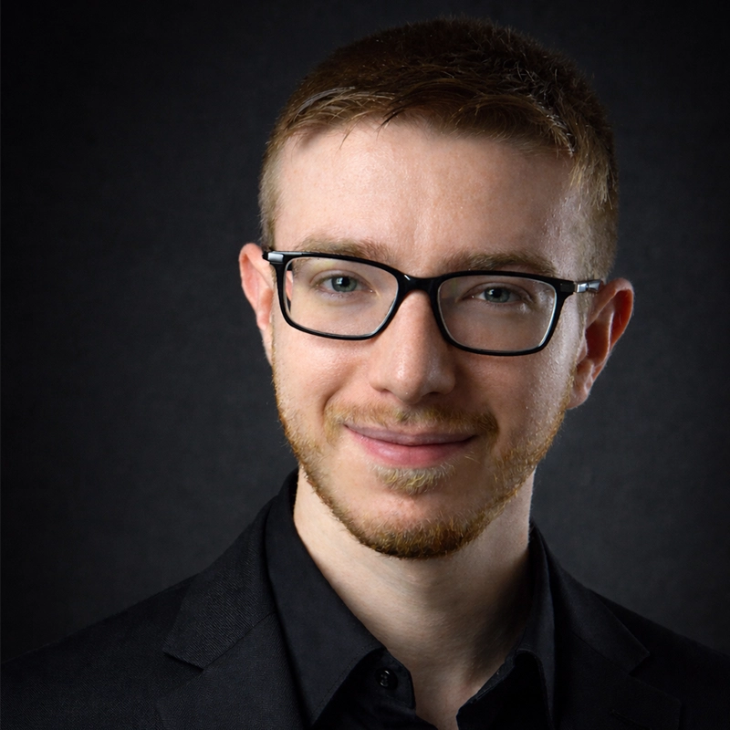 A young man with short light brown hair, trimmed beard, and glasses smiles gently. He wears a black suit jacket and black shirt. The background is plain and dark, making his face and attire stand out clearly.