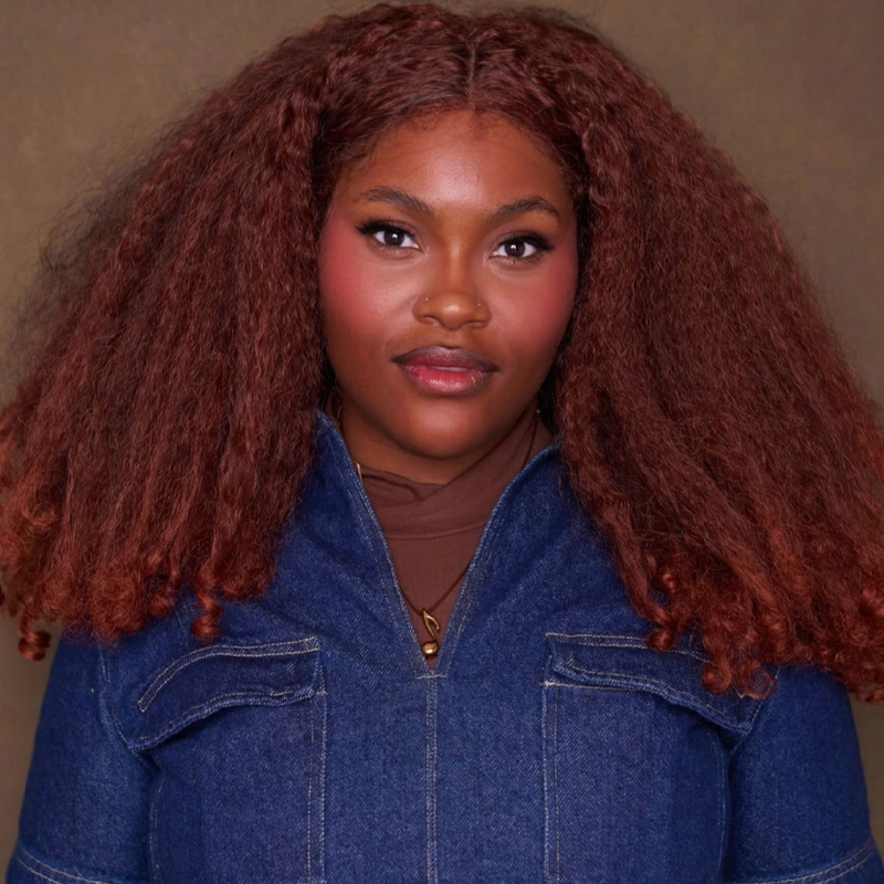 Jana Nicole Prentiss, with voluminous curly reddish-brown hair, wears a blue denim jacket over a brown turtleneck. She has glowing skin, subtle makeup with pink blush and lipstick, and poses confidently against a plain brown background.