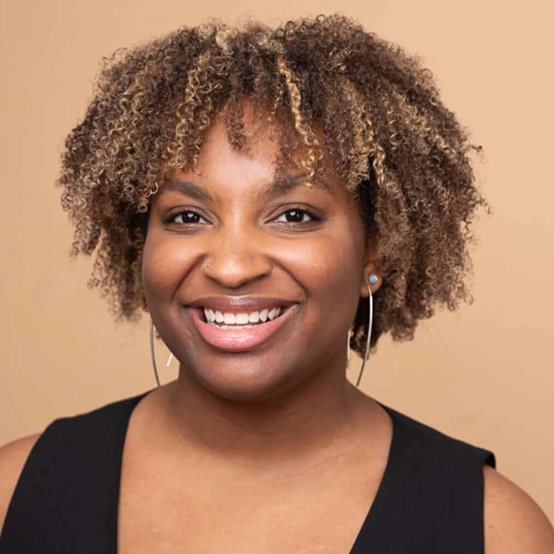 A smiling person with medium brown skin and curly, highlighted hair wears a sleeveless black top and large hoop earrings. Sporting a nose ring and stud earrings, they pose confidently in front of a solid beige background, reminiscent of Christian Clausnitzer’s style.