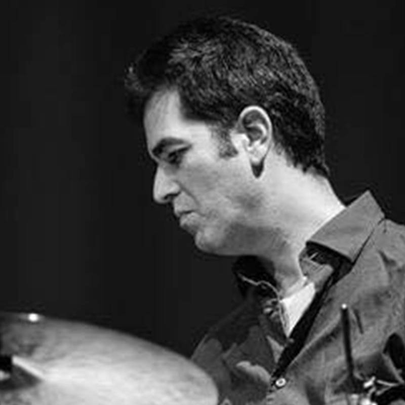 A black-and-white profile shot of Danny Weller with dark hair looking down with focus. He is wearing a buttoned shirt and sits behind a drum set, with only a portion of a metallic cymbal visible in the foreground.