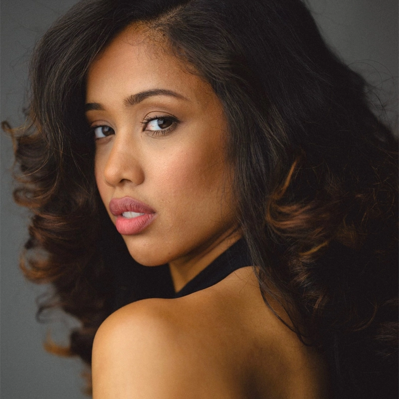 Maya Santiago, with long, wavy dark hair, looks over her bare shoulder toward the camera. She has soft makeup with pink lips and defined brows, wearing a black halter top. The gray background highlights her face and expression.