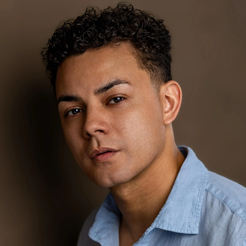 A young man with short, curly dark hair and light brown skin gazes seriously at the camera. He wears a light blue collared shirt and is posed against a plain brown background, in a portrait inspired by Eve Tjoumakaris’s elegant style.