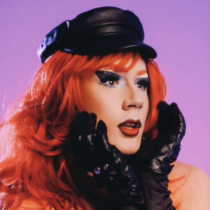 A person with wavy red hair wears a black leather cap and black gloves, holding their face with both hands. Their makeup is dramatic, featuring bold winged eyeliner, red lipstick, and heavy contouring. The background is purple.