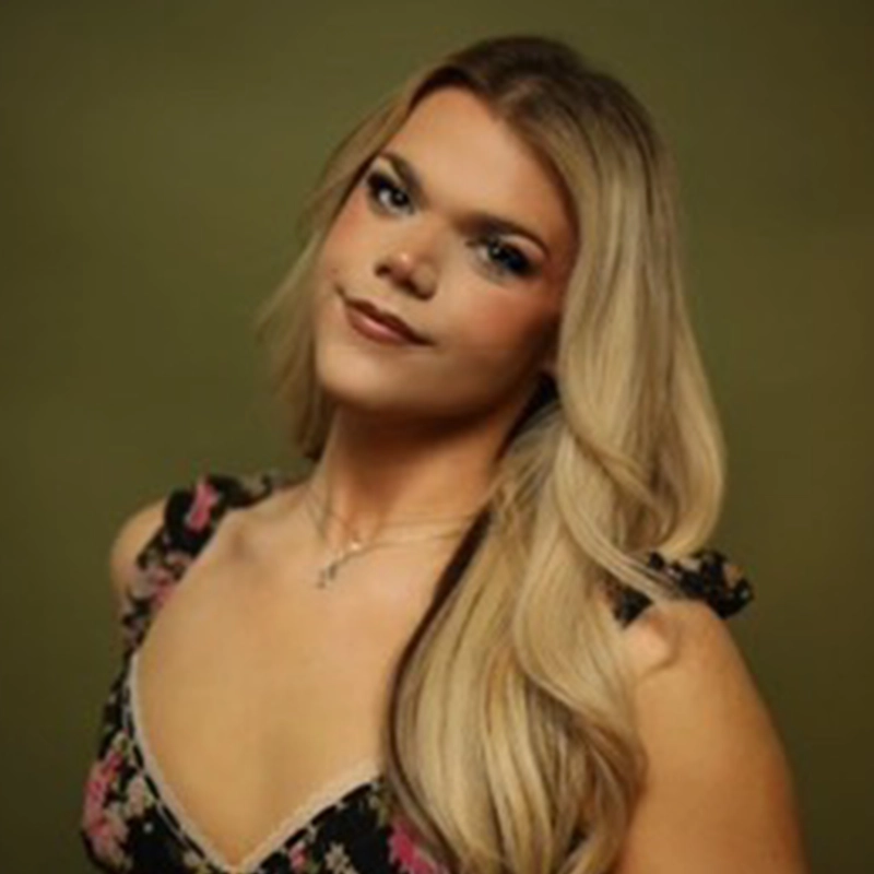 A person with long, wavy blonde hair poses against an olive green background. They wear a black floral dress with ruffled sleeves and a delicate necklace. Their makeup is subtle, with soft eyeshadow and natural lipstick, and they smile gently at the camera.