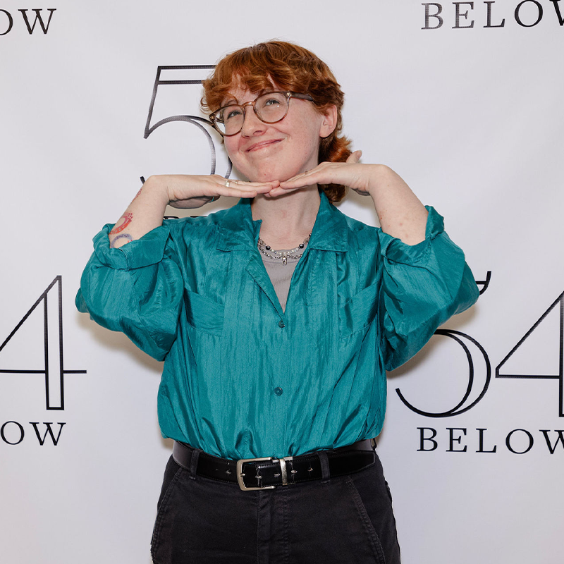 Katie O'Donnell, with short, curly red hair and glasses, smiles with her hands under her chin. She wears a shiny teal shirt and dark pants against a white step-and-repeat banner featuring 