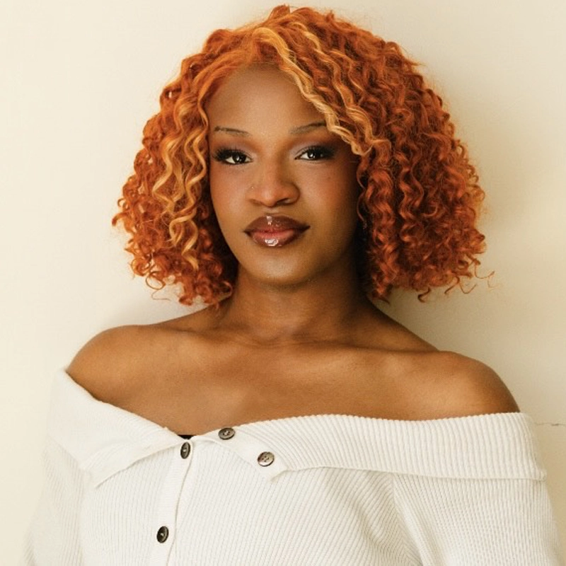 A portrait of Nesziah Dennis, a person with deep skin and vibrant, shoulder-length curly hair streaked with copper and blonde. They are wearing an off-the-shoulder white ribbed top and have a neutral, poised expression against a plain, light background.