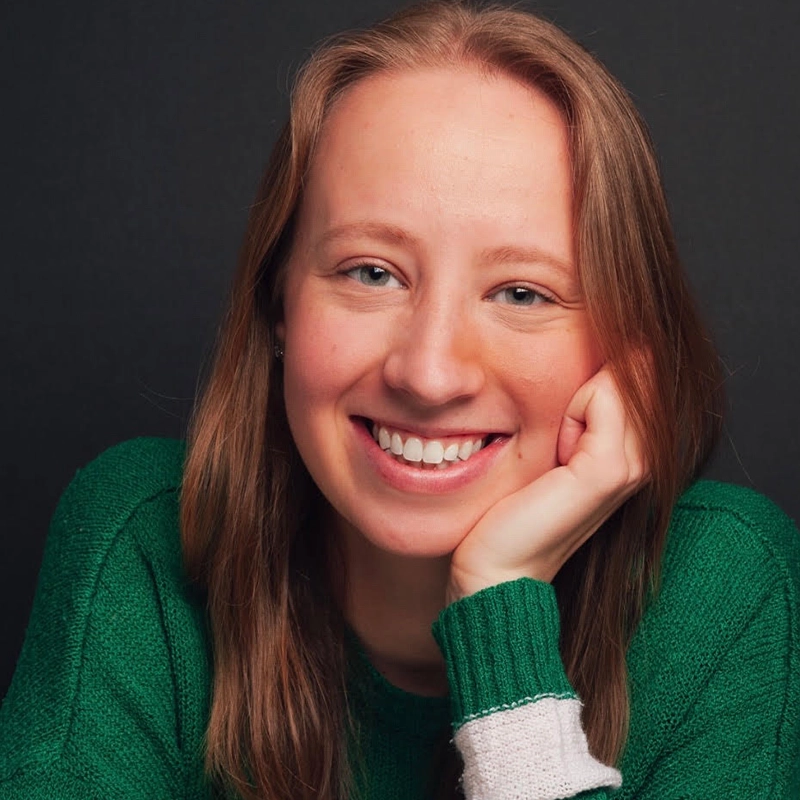 Margot May Waldron, a young woman with straight, light brown hair, smiles warmly. She wears a green sweater with white trim on the sleeve. Her head rests on her left hand. The background is dark, drawing focus to Margot’s cheerful expression and relaxed pose.