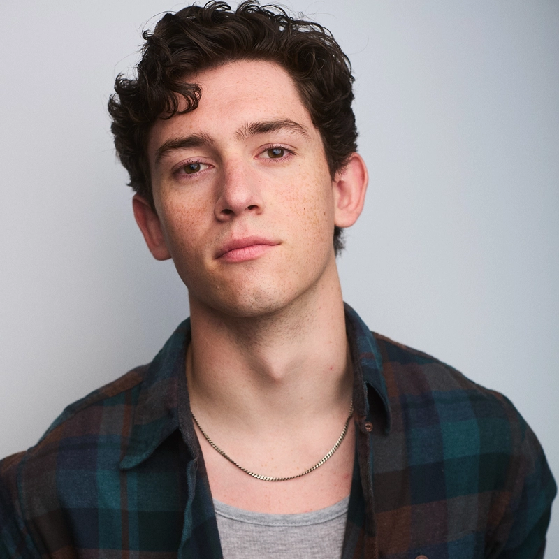 Brayden Bambino, a young man with short curly brown hair and light skin, looks calmly at the camera. He wears a blue and green plaid shirt over a gray T-shirt and a thin silver chain necklace. The background is plain and softly lit.