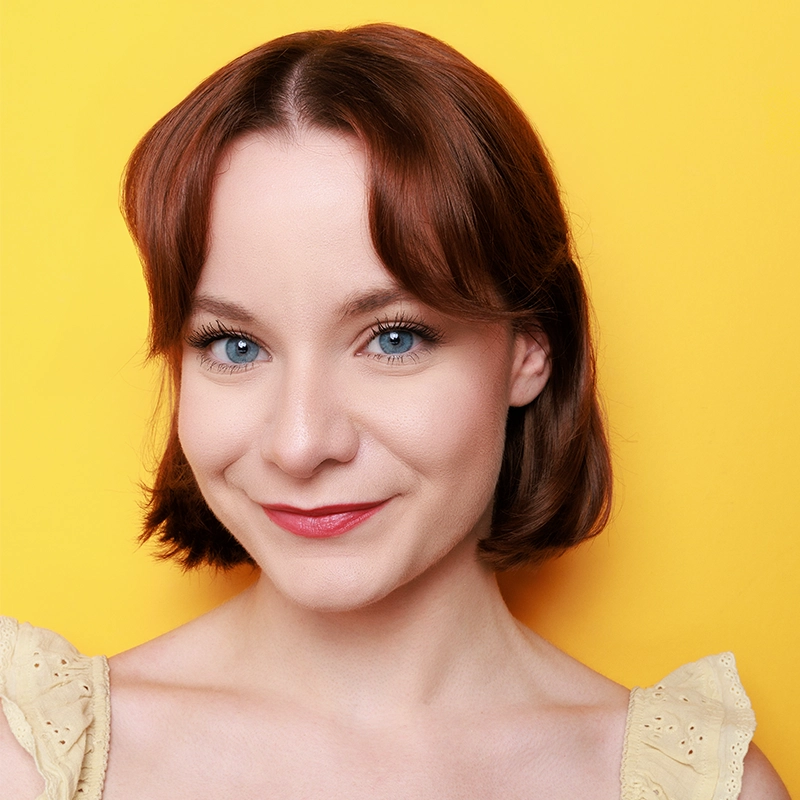 Margot May Waldron, a woman with short auburn hair and blue eyes, smiles softly at the camera. She wears a light yellow lace top with ruffled sleeves. The background is solid bright yellow, creating a cheerful, vibrant atmosphere. Her makeup is natural with pink lipstick.