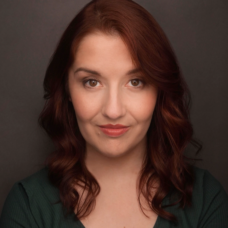 A woman with fair skin and wavy auburn hair is facing the camera, wearing a dark green top. Margot May Waldron has neutral makeup, a gentle expression, and slightly smiling lips. The background is plain and dark, emphasizing her face. The lighting is soft and even.