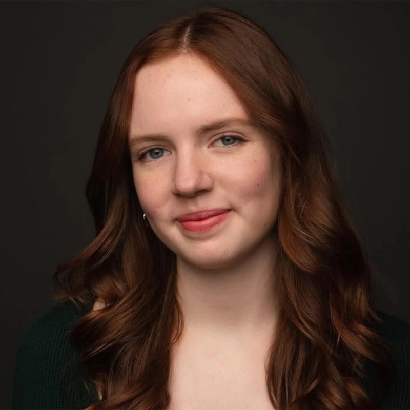 A young woman with long, wavy auburn hair smiles softly at the camera. She has fair skin, light blue eyes, and is wearing a dark green top. The background is plain and dark, highlighting her face and hair.