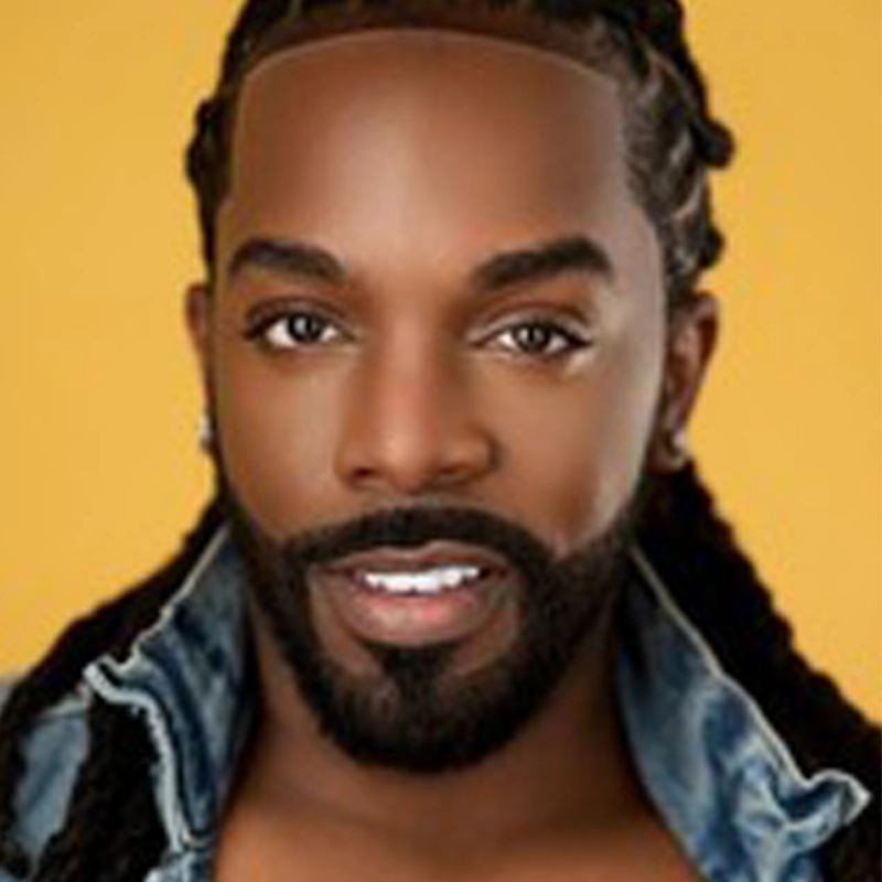 A confident Black man with neatly groomed beard and mustache, long braids, and diamond stud earrings poses against a vibrant yellow background. Channeling Sherrod Brown’s effortless cool, he wears a denim jacket off his shoulders and smiles subtly at the camera.
