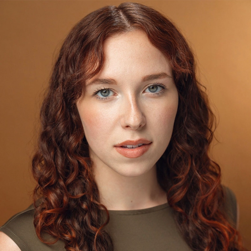 A woman with fair skin, long wavy auburn hair, and blue eyes looks at the camera with a neutral expression. She is wearing a sleeveless olive-green top. The background is a warm, solid brown color. The lighting is soft and even, highlighting the features of Margot May Waldron.