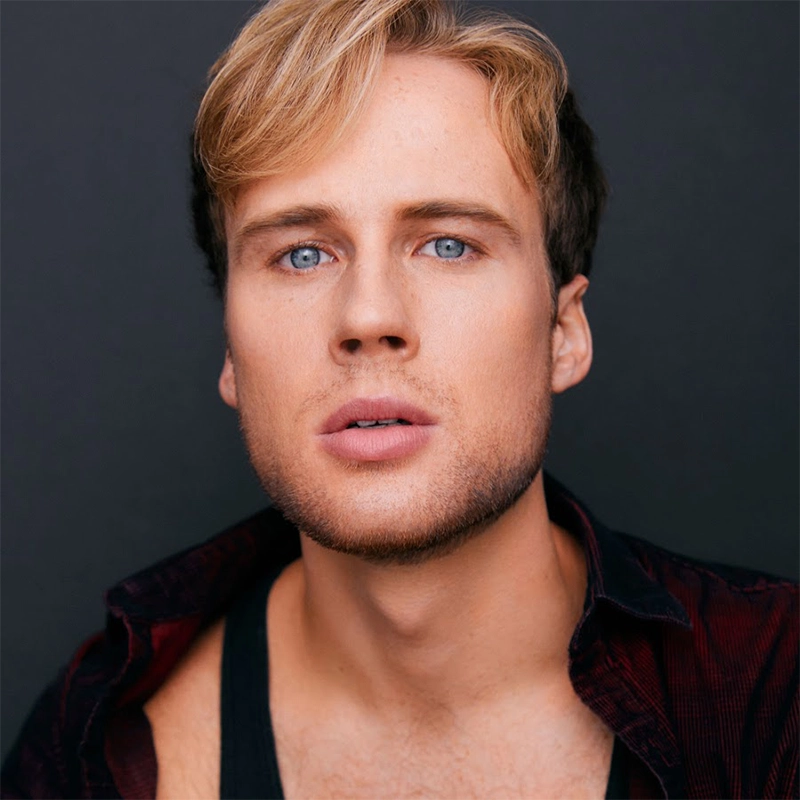 A young man with light skin, tousled blond hair, and blue eyes gazes directly into the camera with a neutral expression. Charlie Tingen wears a dark open-collared shirt over a black top against a plain dark background. His face is well-lit and clean-shaven.