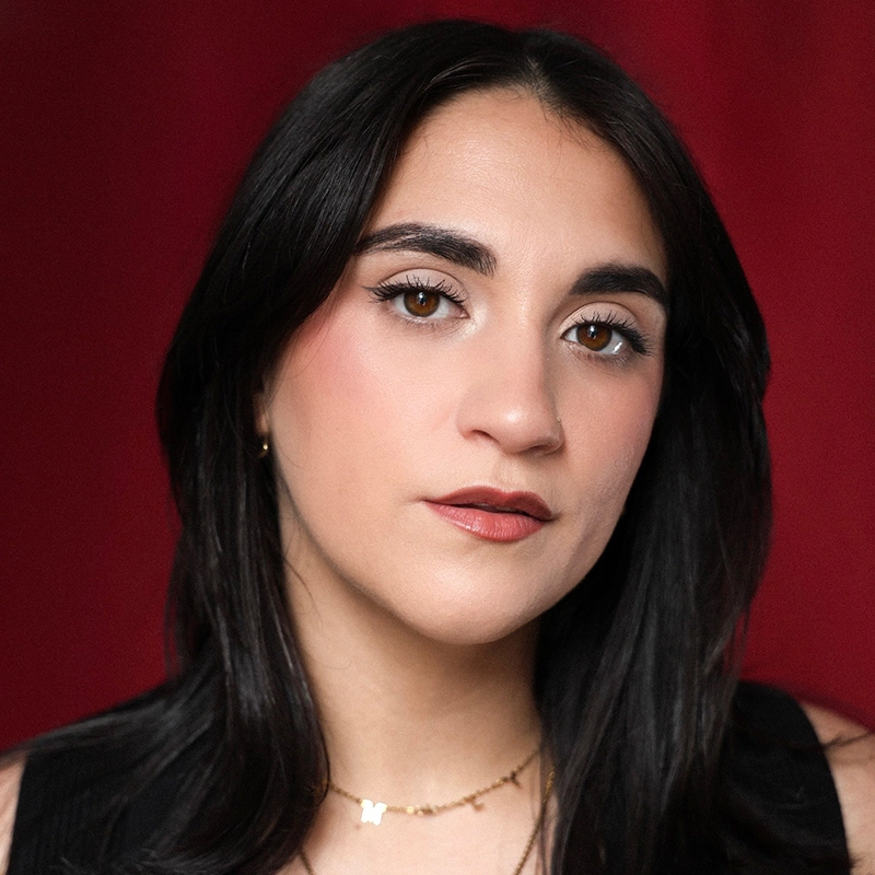 Samantha Gibbs, a woman with long, dark hair and brown eyes, looks at the camera with a neutral expression. She wears subtle makeup, a gold 