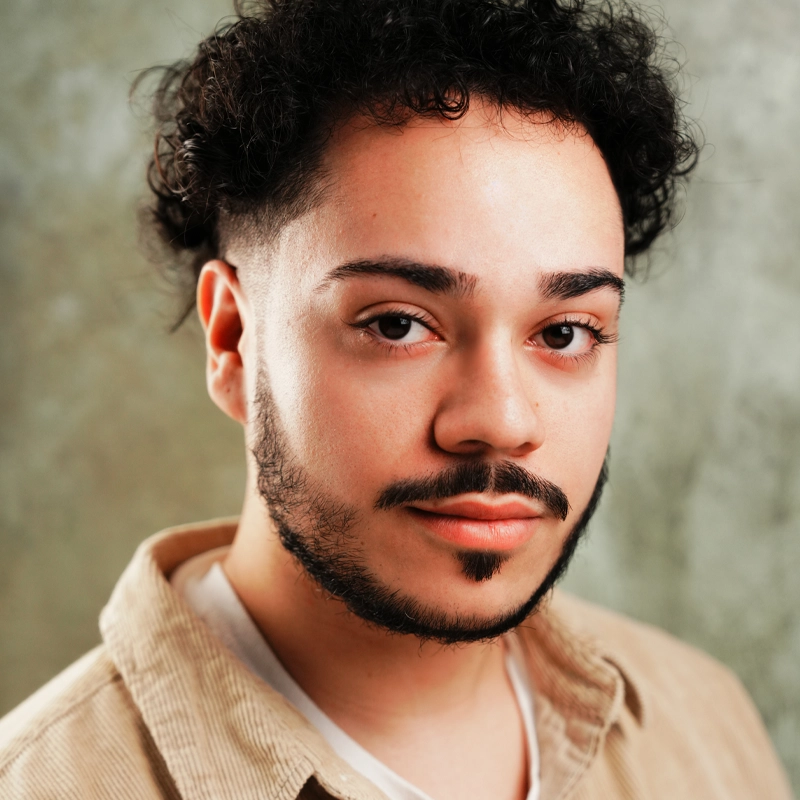 Jeremy Fuentes, a young person with short curly dark hair, shaped facial hair, and neatly groomed eyebrows, looks into the camera with a neutral expression. They wear a cream corduroy shirt over a white top against a softly blurred green-gray background.