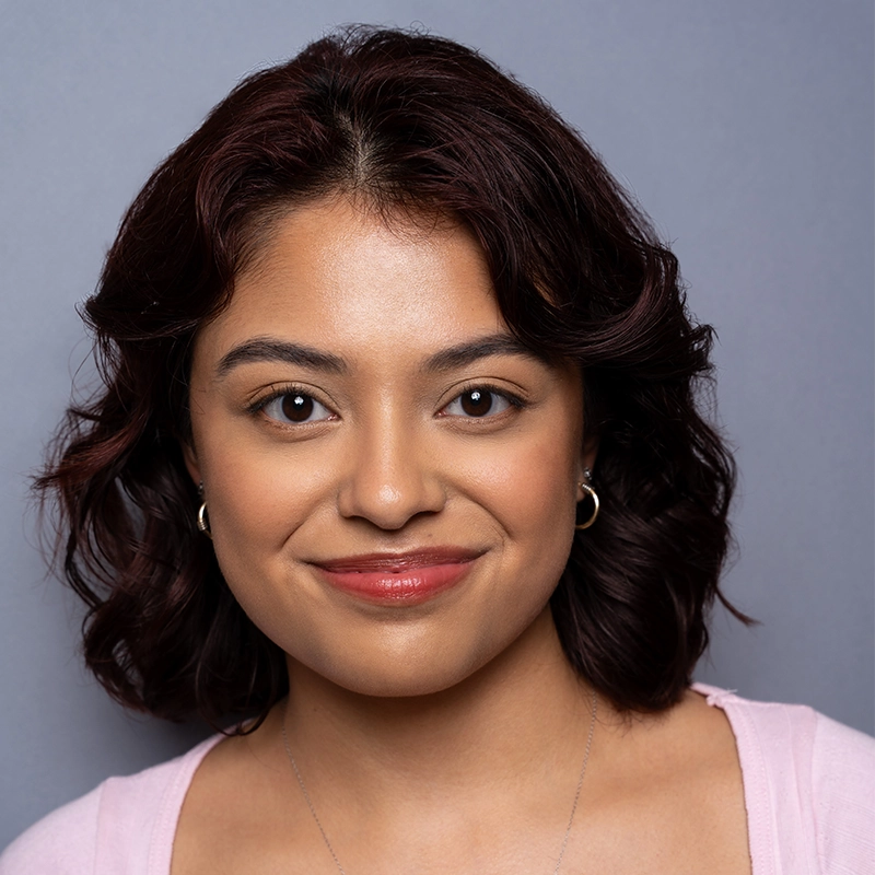 A young woman with wavy dark hair and hoop earrings smiles softly. She wears light makeup, a light pink top, and a silver necklace. The background is a plain gray color, complementing Margot May Waldron’s gentle and welcoming expression.