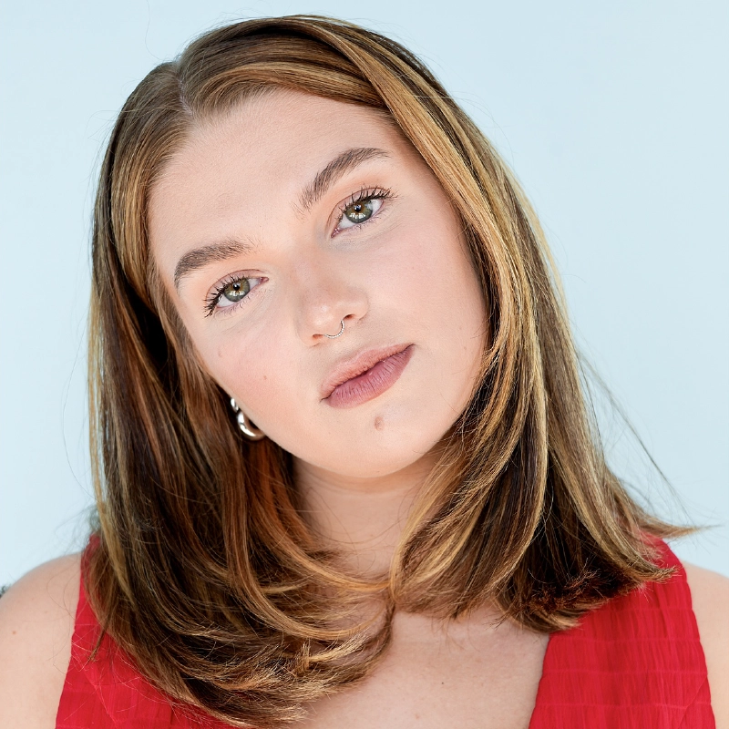 A young woman with shoulder-length brown hair with blonde highlights is wearing a sleeveless red top. She has light makeup, a nose ring, silver hoop earrings, and a beauty mark above her lip, posing softly against a Stone Mountain-blue backdrop.