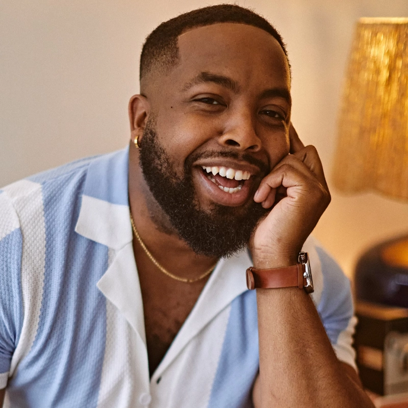 A smiling man, Manny Houston, with a neatly trimmed beard and short hair rests his head on his hand. He wears a pale blue and white shirt, gold necklace, small hoop earring, and brown watch. Warm lighting and a lamp in the background create a cozy atmosphere.