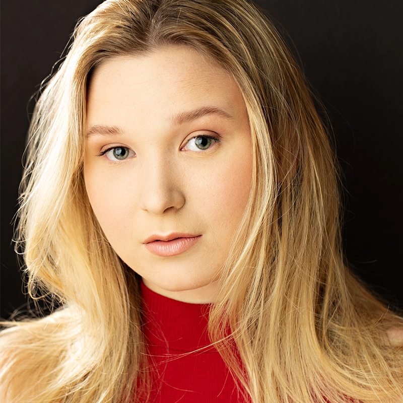 A young woman with fair skin and long, straight blonde hair looks at the camera. She wears a sleeveless red top and has a neutral expression. The background is black, and soft lighting highlights her facial features and hair, giving Margot May Waldron an elegant, striking presence.