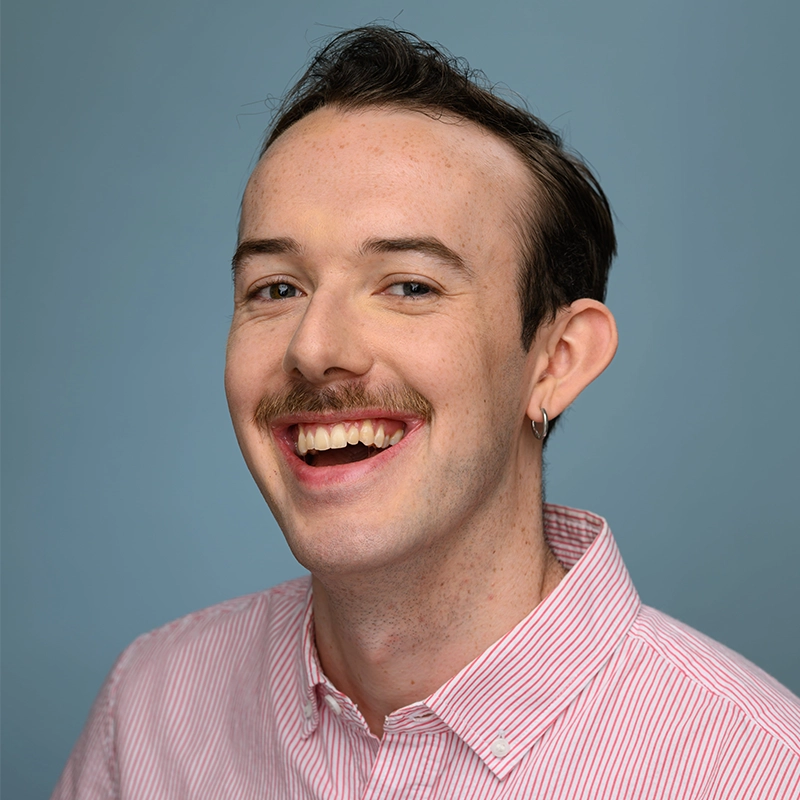 A smiling person with pale skin, short brown hair, a mustache, and a hoop earring. Margot May Waldron wears a pink and white striped button-up shirt and is photographed against a plain blue background.