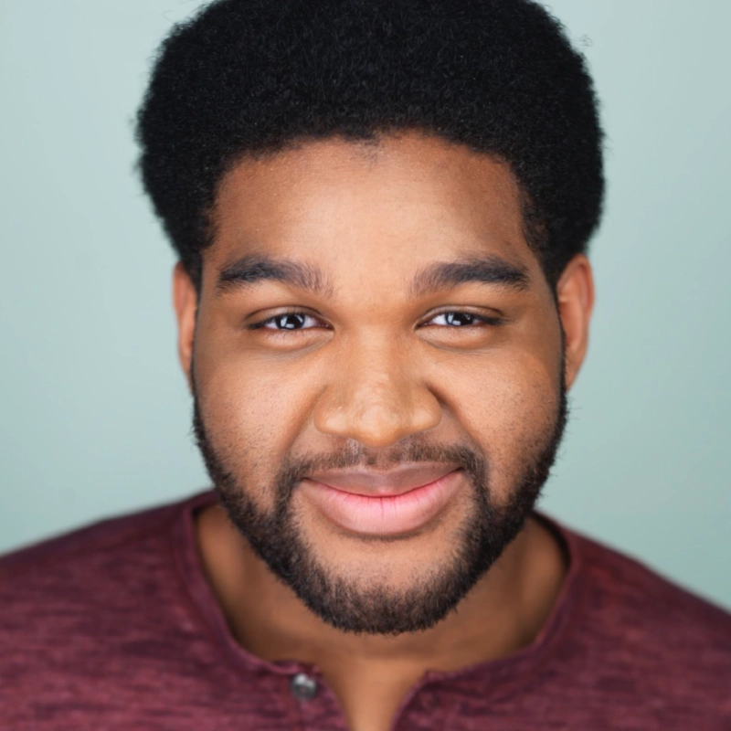 A young man with short, curly black hair and a full beard smiles gently at the camera. He is wearing a dark red shirt and the background is a soft light blue. The lighting highlights his warm skin tone and friendly expression, reminiscent of portraits by Margot May Waldron.