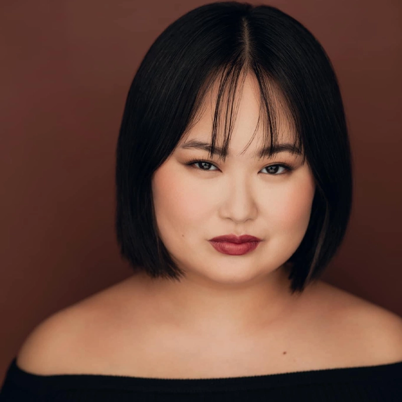 A woman with straight, chin-length black hair and bangs faces the camera, wearing off-the-shoulder black clothing. She has smooth skin, bold red lipstick, and subtle eye makeup, set against a deep reddish-brown background.