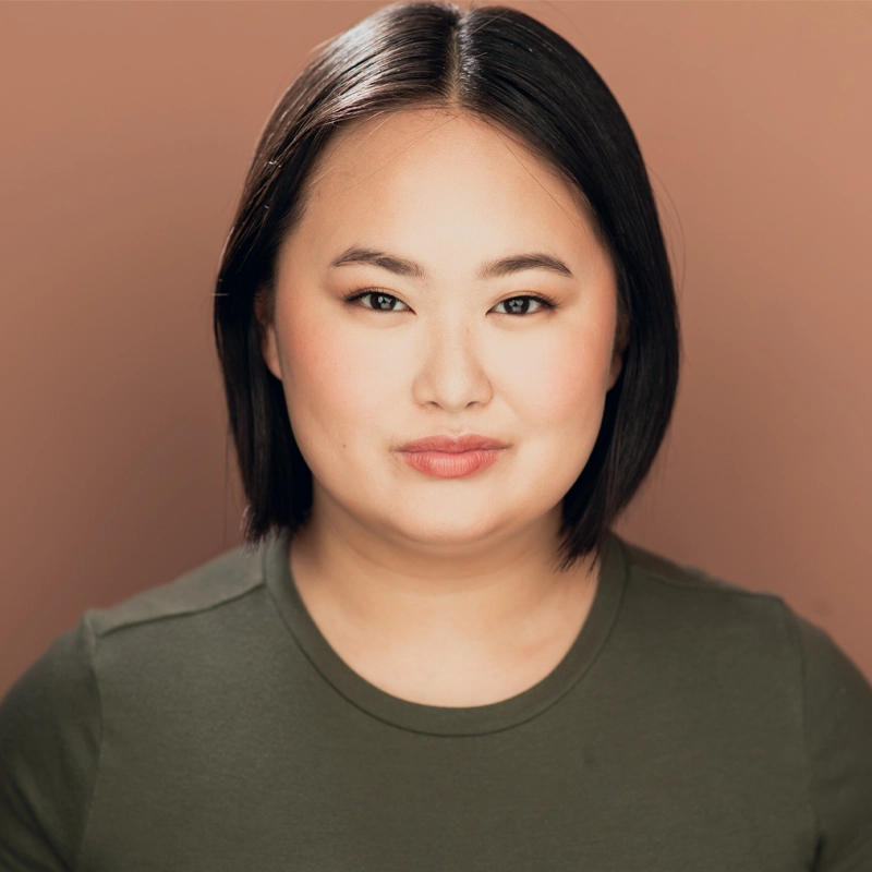 A woman with straight, chin-length dark hair and light skin smiles softly. She wears a green crew-neck shirt and neutral makeup. The background is a solid, warm brown, and the lighting is even, highlighting her facial features.