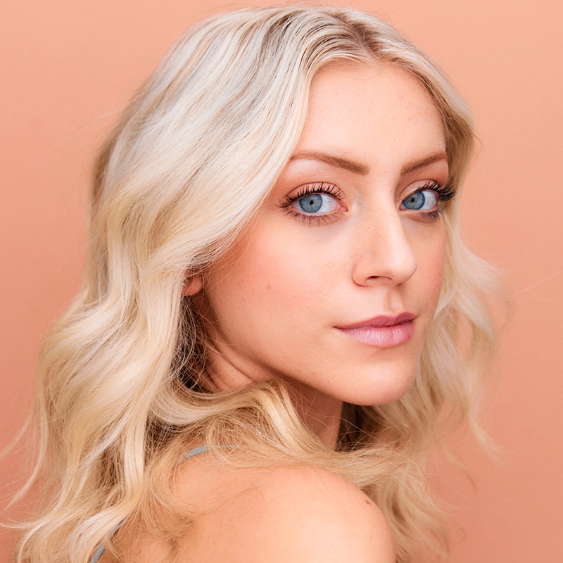 A young woman, Margot May Waldron, with wavy platinum blonde hair and bright blue eyes gazes over her shoulder at the camera. She has even fair skin, soft pink lips, and subtle makeup. The background is a peachy beige color.