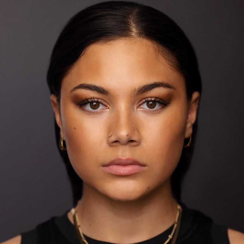 A young woman with medium skin tone, straight dark hair, and brown eyes looks directly at the camera. She wears gold hoop earrings, layered necklaces, and a nose ring. Her expression is neutral, and the background is plain and dark.