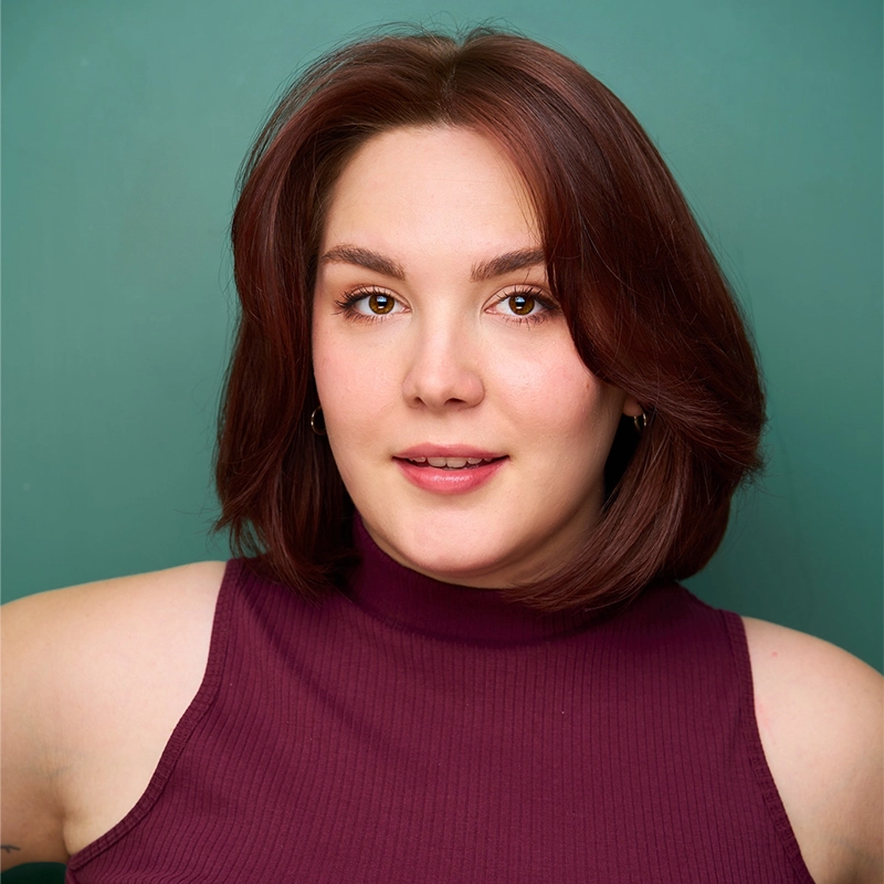 A young woman with fair skin and shoulder-length brown hair poses against a teal background, her style as striking as Stone Mountain. She’s wearing a sleeveless, burgundy mock-neck top, subtle makeup, and offers a slight smile with direct eye contact.