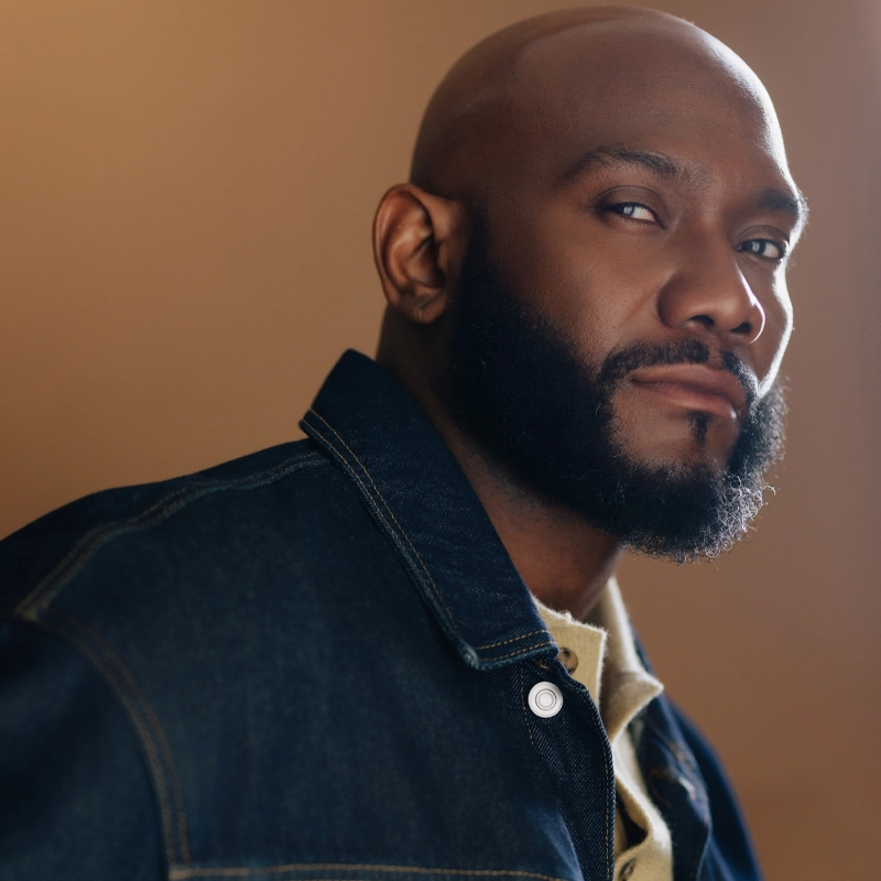 Apollo Levine, a bald man with a full beard, gazes confidently at the camera. He wears a dark denim jacket over a light-colored shirt. The warm brown background and soft lighting highlight his facial features.