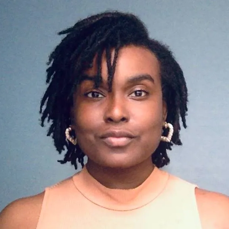 A person with short, textured black hair wears large, twisted hoop earrings and a sleeveless, peach-colored top. Manny Houston faces the camera against a plain blue background, wearing a neutral expression.