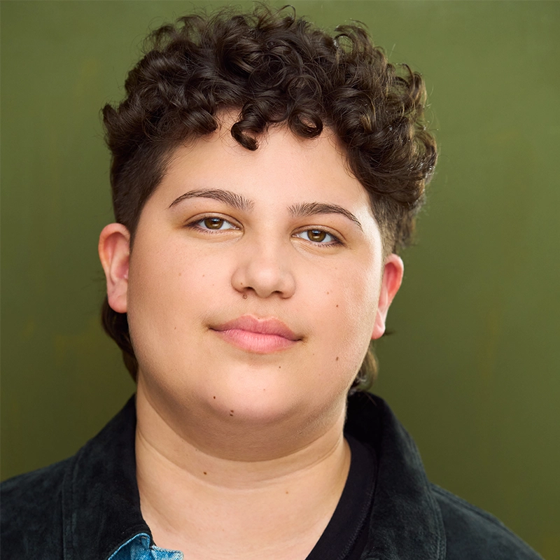 A person with short, dark, curly hair and light skin looks directly at the camera with a calm expression. They are wearing a dark jacket over a light blue shirt, set against a solid olive green background reminiscent of Stone Mountain’s earthy tones.