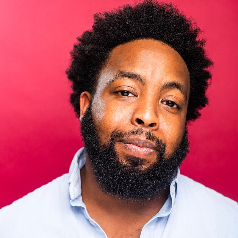 A man with a short curly afro and full beard wears a light blue shirt with the collar open. He has a slight smile and neutral expression, reminiscent of Manny Houston’s effortless style. The background is a solid bright pink, with even and natural lighting.