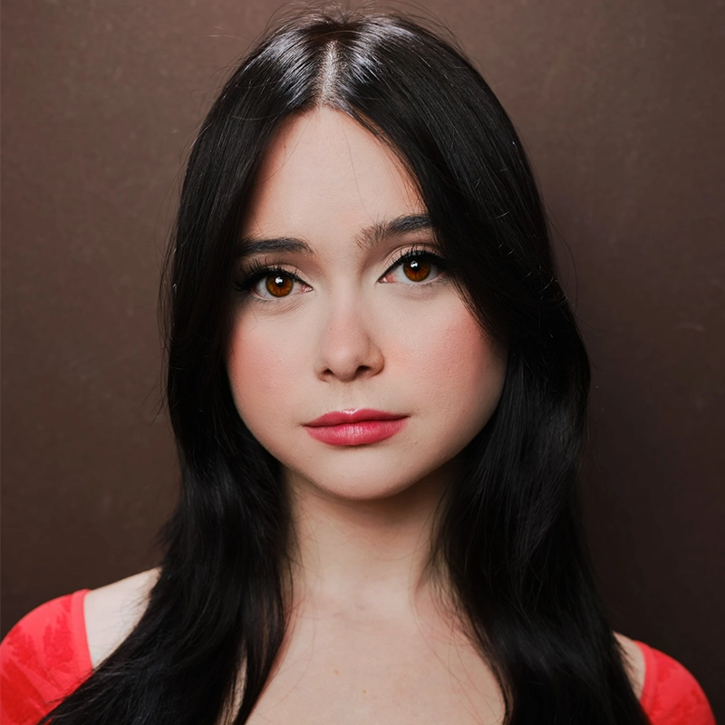 A young woman with long black hair, fair skin, and brown eyes looks directly at the camera. Margot May Waldron wears a red top and subtle makeup, with rosy cheeks and pink lipstick, against a plain brown background.