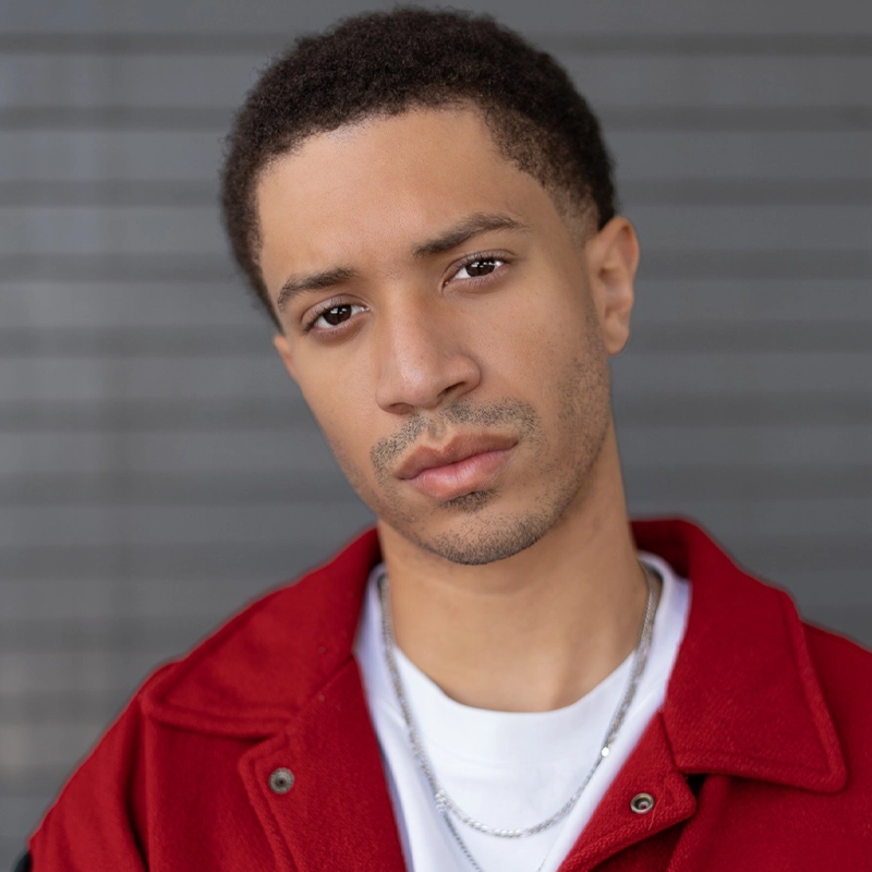 Matte Martinez, a young man with short curly hair, wears a red jacket over a white shirt and silver chain necklaces. He gazes seriously at the camera against a gray background with horizontal lines, his expression neutral in the soft light.