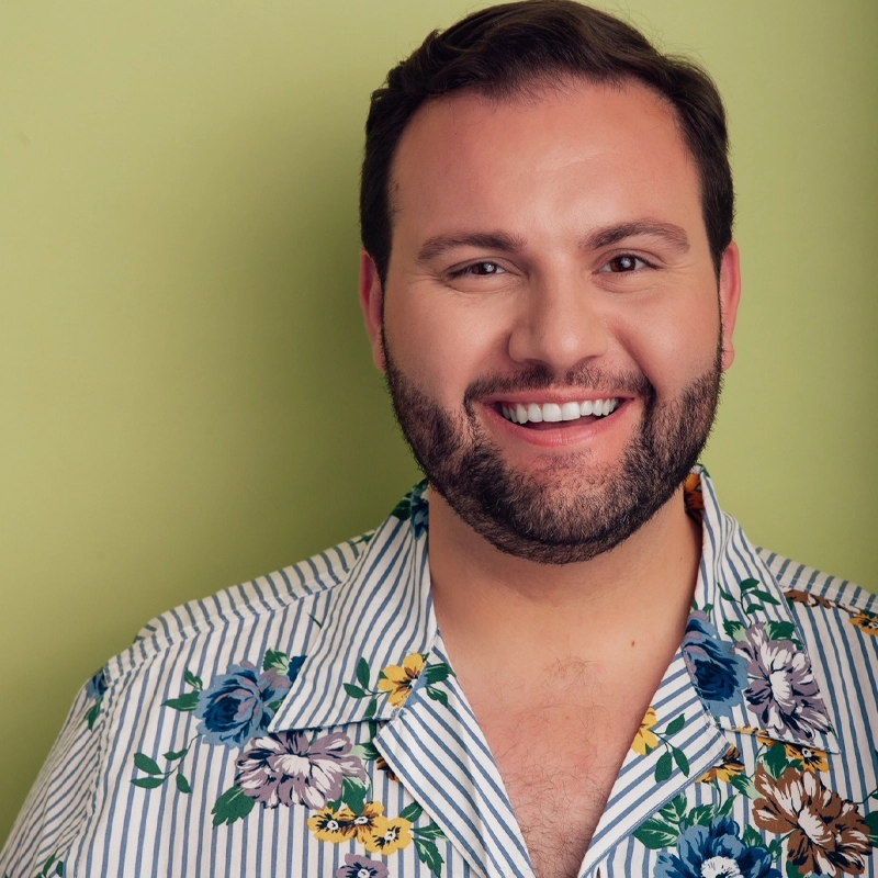 A smiling man, Mitchell Turner, with short dark hair and a beard stands in front of a light green background. He wears a white shirt with blue vertical stripes and a colorful floral pattern, facing the camera, looking cheerful and friendly.