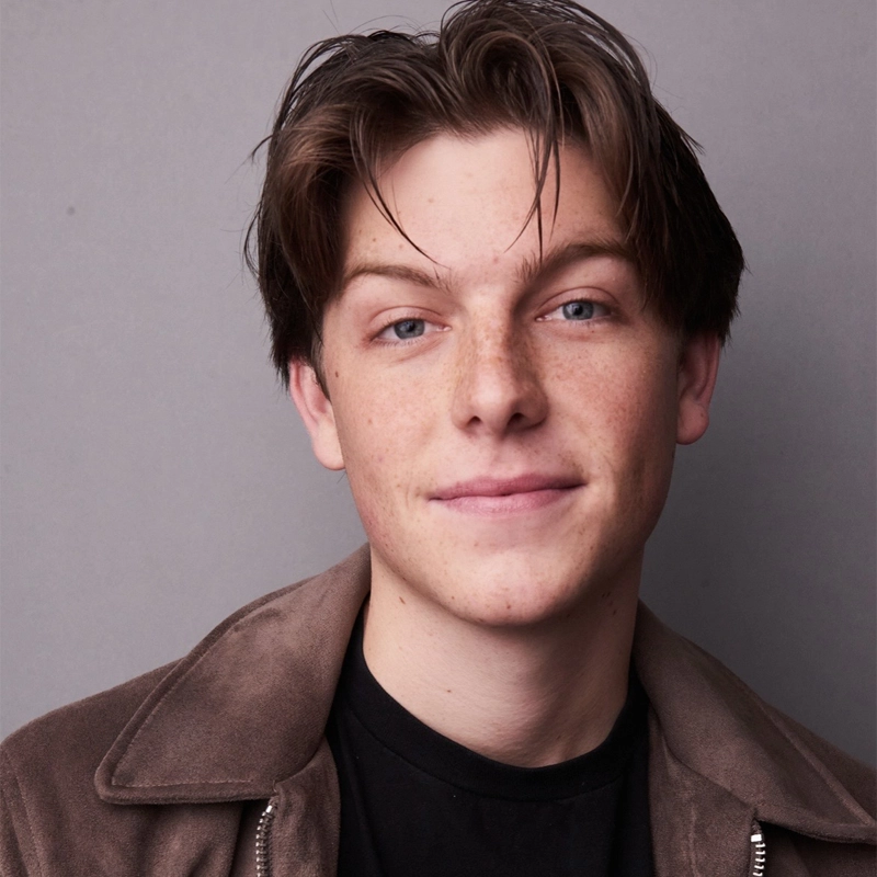 A young person with fair skin, blue eyes, and medium brown hair parted down the middle smiles gently. Michael Moretti wears a brown jacket over a black shirt and stands against a plain gray background.