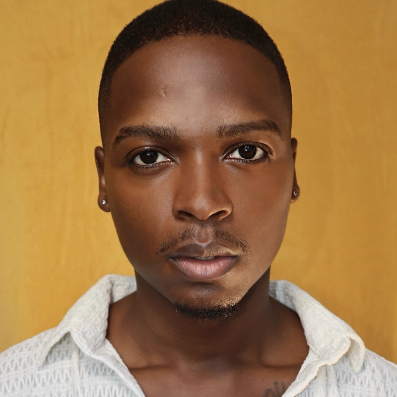 A young Black man, Aramie Payton, with short hair and a trimmed goatee gazes directly at the camera. He wears small stud earrings and a white textured shirt with the collar open, set against a solid mustard yellow background for vibrant contrast.