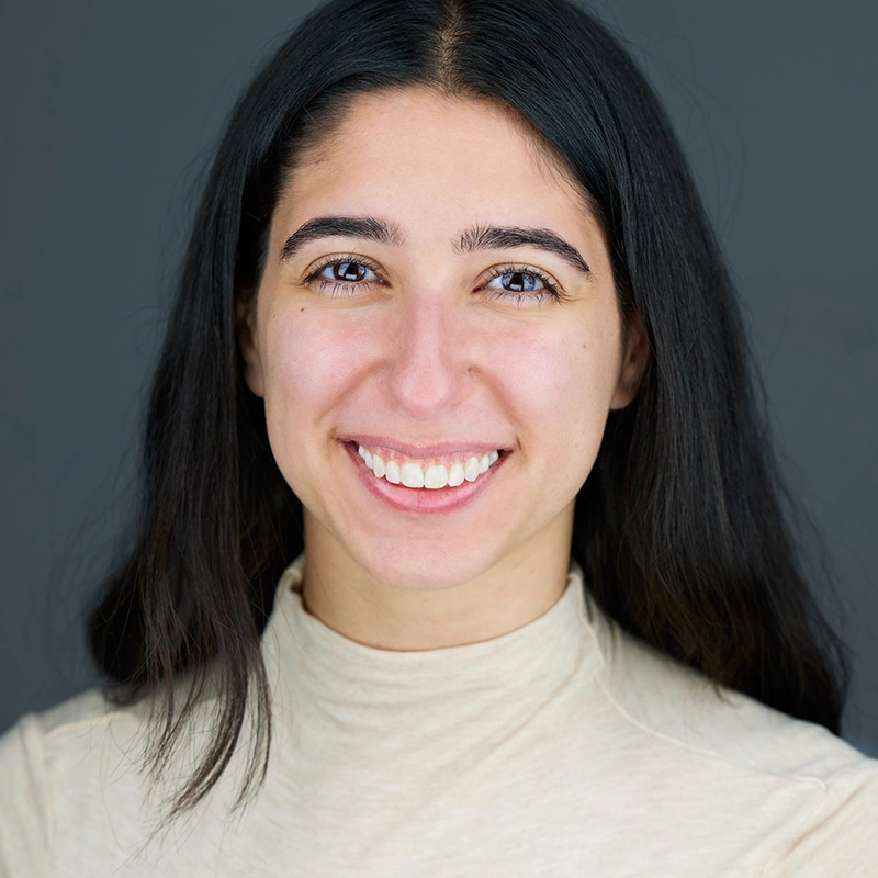 Olivia Berkson, a young woman with long dark hair, smiles at the camera in a light beige turtleneck top. The plain dark gray background creates a professional and friendly portrait atmosphere.