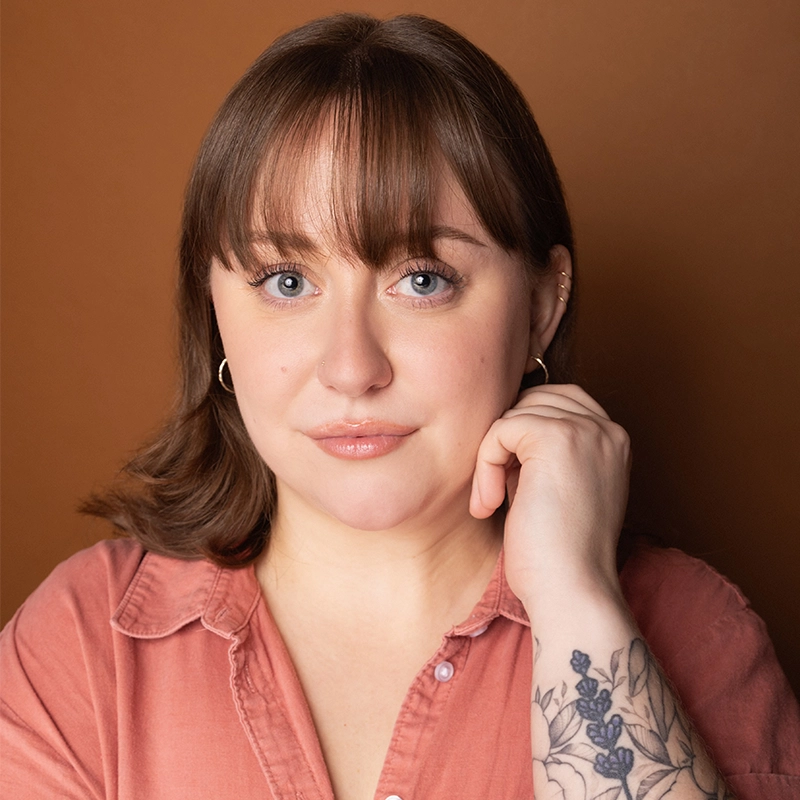 A woman with light skin, straight brown hair with bangs, and blue eyes poses against a brown background. She wears a peach-colored shirt, small hoop earrings, and has a floral tattoo on her left forearm—her style reminiscent of Stone Mountain chic.