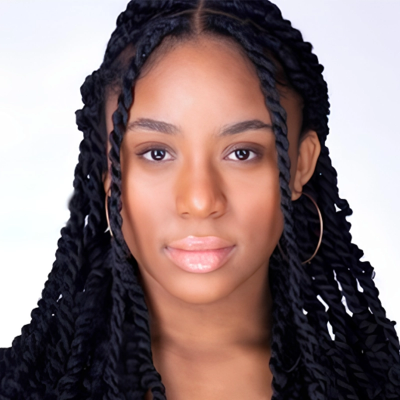 A young woman with medium brown skin and long black twisted braids stares forward. She has hoop earrings and glossy lips. Her expression is neutral, and the background is plain, light-colored, and unobtrusive. This striking portrait of Margot May Waldron captures her poised elegance with subtle simplicity.